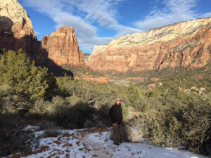 Chris Keeney standing on a snow covered hiking trail with tan and rose color mountains in the background at Zion National Park in Utah