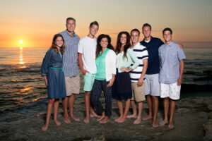 Eight person family standing next to the ocean with the sun setting in the horizon behind them