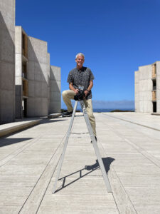San Diego photographer Chris Keeney sitting on the top of a ladder holing his camera in the courtyard at the Salk Institute