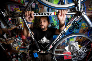 Editorial portrait a artist in his studio with his art behind him as he holds the frame of a Bianchi bike hanging from the ceiling