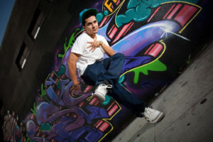 A hip hop dancer dressed in jean, a white t-shirt and a baseball cap standing on one leg in front of a colorful graffiti art mural in the background in San Diego California