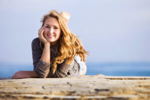 blonde teenage girl lying on the bluff with her hand on her face and smiling at the camera and the ocean behind her