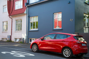 Red car parked on the street in front of a dark gray house with the number 47 on the side reflecting a red house that is across the street in the window. There is a large number 30 painted on the street.