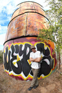 DJI Mini 3 Pro 360 degree panoramic drone photograph taken in Santa Barbara, California of San Diego photographer Chris Keeney standing in front of an old rusted water tower with graffiti painted on the side of it. Chris is wearing green pants, a white hiking shirt and a green bucket hat.