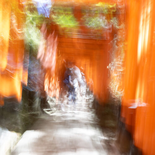 an experimental photograph of Fushimi Inari Shrine that has the feeling of moving through time with warm tones of yellow and orange accented with green, black and tan colors. A path with a person in the distance can be seen in an artistic way