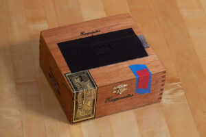 Color photograph of a Arturo Fuente Exquisitos wooden cigar box homemade pinhole camera with a black chipboard paper shutter on a wooden countertop.