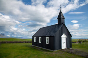 A small wooden black church with white multiple paned windows and a white door. located in a wide open landscape with green grass and the ocean and mountains in the background.