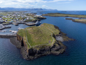 DJI Mini 3 Pro drone photograph of Súgandisey Island Lighthouse in Stykkishólmur, Iceland. You can see the Iceland Coast Guard ship in the background named ICGV Þór (Thor) which is an UT 512L type offshore patrol vessel. The sky is blue with mountains and small islands in the distance.