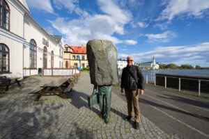 Photo of Chris Keeney standing next to the Monument to the Unknown Bureaucrat statue next to the City Hall building with blue sky and puffy white clouds in Reykjavik, Iceland 2023