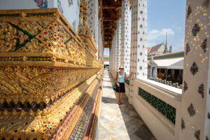 A view of a woman standing in a hallway with thousands of tiny colored mirrors that reflect the sunlight onto the wall and people walking next to them at The Grand Palace in Bangkok Thailand.