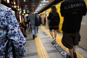 A photo of people walking to the subway train in downtown Tokyo, Japan. A young man can be seen waring a black ALLSAINTS shit and the girl in front of him is wearing a black skirt, sneakers and black and gold trimmed jacket. A woman in the foreground is wearing a purple and light blue spotted white jacket. There is a lot of yellow painted lines and walls in the station.