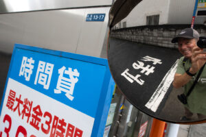 A reflection of Chris Keeney smiling at the camera in a round concave mirror on the street in Tokyo, Japan. Japanese kanji can be seen on a blue and white sign and on the black asphalt of the street.