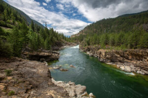 looking up stream at the Kootenai river in Lincoln County Montana. The water is teal green the sky is blue with white and gray clouds and there's a forest of evergreen trees on either side of the river.