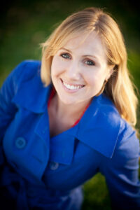 Head and shoulder headshot portrait of a blonde woman with blue eyes wearing a blue coat and red shirt smiling at the camera