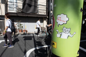 A people walking and riding a bicycle on the street in Tokyo Japan during the daytime. You can see a cat with Japanese kanji that with a text bubble saying "No, it's dangerous"