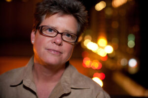Head and shoulder portrait of a short hair woman wearing glasses and a tan shirt with city lights behind her in downtown San Diego, California