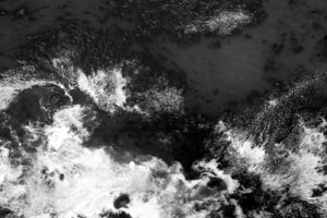 Black and white photograph of a view of the ocean from a plane window banking so the you're looking straight down at the ocean. You can see the white wash of the waves breaking and kelp floating on the surface of the water