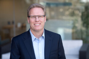 color photograph of a business man in a blue suit jacket and button down shirt wearing glasses and smiling at the camera. Soft out of focus glass office window in the background