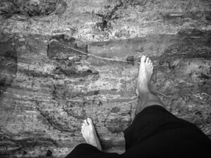 black and white photo showing Chris Keeney's legs and feet standing on top of a large flat granite rock with lines and textures in it. Chris is wearing black shorts and is barefoot in the photo