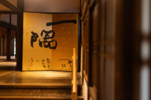 a color photograph of one of the hallways in the the Shōren-in Buddhist temple in Kyoto Japan. You can see a large work of art with Japanese brush stroke kanji characters written on a gold two panel wood art piece.