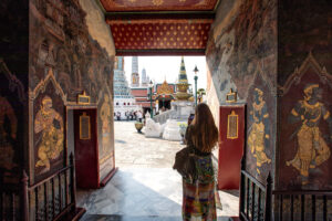 wide angle color photograph of a women stain in an entryway looking out onto a plaza with multiple stupas. It appears she's taking a photo with her phone of a person standing on a staircase in the background.
