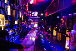 a photo of the inside of a dark bar lit with red and blue lighting showing people seated at the bar and small tables