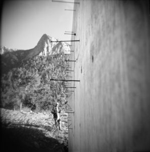 black and white photograph of a man standing next to a concrete wall with nails coming out of it and in the background is a large granite peak called Tahquitz Peak in idyllwild, California