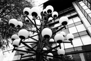 black and white geometrically composed street photograph of a city light shaped like a tree with multiple white light globes with a building with square windows behind it