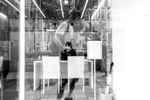 Black and white photo of a reflection of Chris Keeney in a store window in Japan. You can see a Japanese woman working at a desk inside.