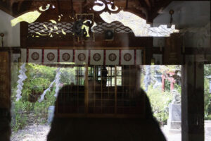 a reflection of a person looking into a window of a Shinto shrine in Hachinohe Japan. You can see a torii gate, a tomoe, a shade with shimenawa and a portrait hung on the wall.
