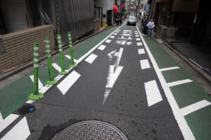 a view looking down a one way street in Asakusa, Tokyo Japan with and green directional graphics painted on the road. People are walking on the side and car is driving down the street in the distance towards the camera.