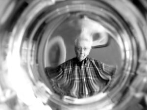 Black and white photo of an older gentleman wearing a plaid button down shirt and glasses looking into the camera. It appears as if the photographer has the camera in a glass shooting through the top. The image is distorted on the edges and clear in the circular center.