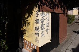 Color photograph of the entry way to a fortune tellers location that has a sign hand written in Japanese kanji that reads Four Pillars of Destiny and an illustration of a palm with lines on it