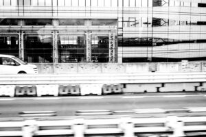 Black and white photograph of a road next to some buildings that have vertical and horizontal lines and windows. A car is seen driving on the left hand side of image. Tokyo Japan 2025
