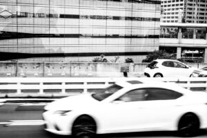 Black and white photograph of a road next to some buildings that have vertical and horizontal lines and windows. Cars are seen driving both direction of street. Tokyo Japan 2025