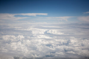 color photograph of blue sky in the horizon and a blanket of soft white fluffy clouds in the foreground. photographed from an airplane window.