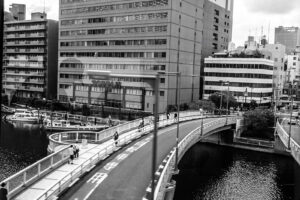 Black and white photography of a curved bridge over a waterway in downtown Tokyo Japan showing people walking, cars on the street and boats docked next to large business buildings.