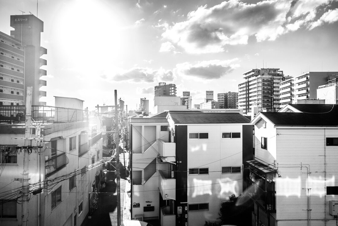 Black and white photograph of the city of Osaka through a window in a passing train. The afternoon setting sun shines at the train windows and they reflect sunlight onto the buildings creating motion picture like frames of light.