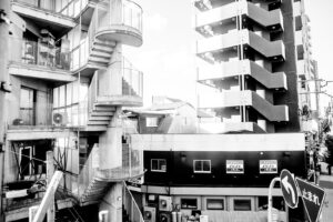 black and white street photography Osaka Japan showing apartment building with outside staircases, a sidewalk with a person walking on it.