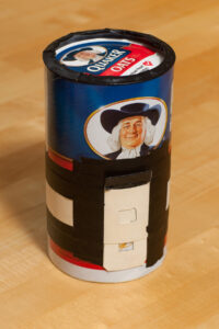 color photograph of a Quaker Oats oatmeal can that has been converted into a pinhole (lensless) no lens camera. There is a homemade sliding paper shutter on the front and black photo tape keeping it in place