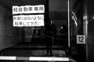 A simple black and white photo of Chris Keeney's reflection in a window of a store at night in Kyoto, Japan with Japanese kanji written in two white rectangles a stripped metal bar on the bottom and the number 12 on the right side of the image. Tile floor and stone wall with a reflection of buildings in the background. Streaks of light on the window.
