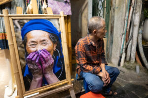 A man sits in a Hanoi alleyway next to a framed "Hidden Smile" portrait by Réhahn, featuring Madame Xong with indigo-stained purple hands.