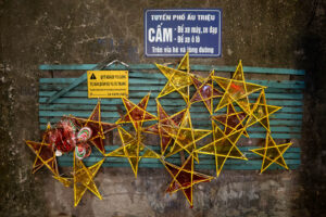 Handmade yellow and red star lanterns hanging on blue wooden slats against a textured wall with Vietnamese street signs in Hanoi's Old Quarter during Tet.